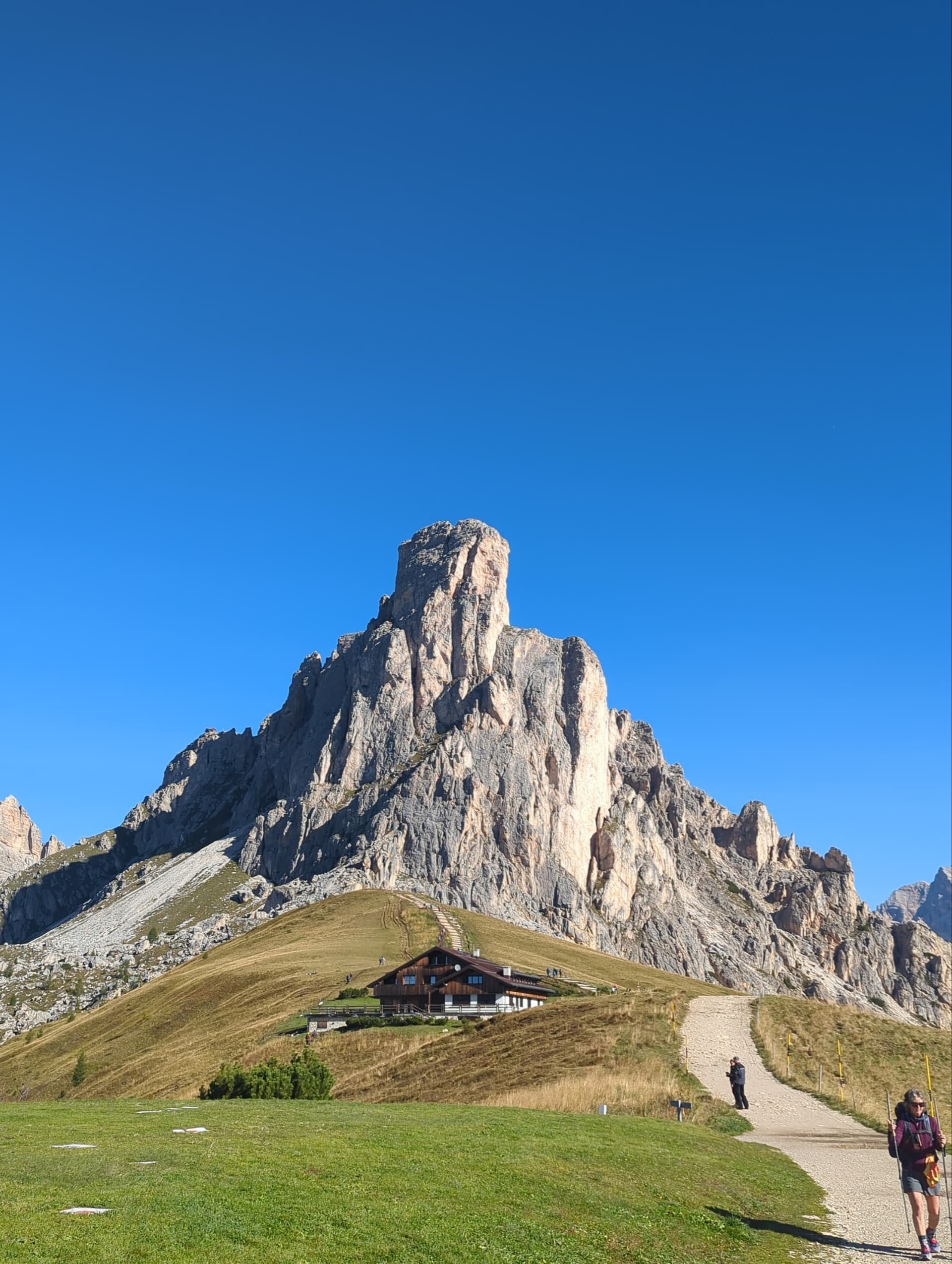 Hikers on a trail leading toward a mountain hut below a jagged peak under a clear blue sky