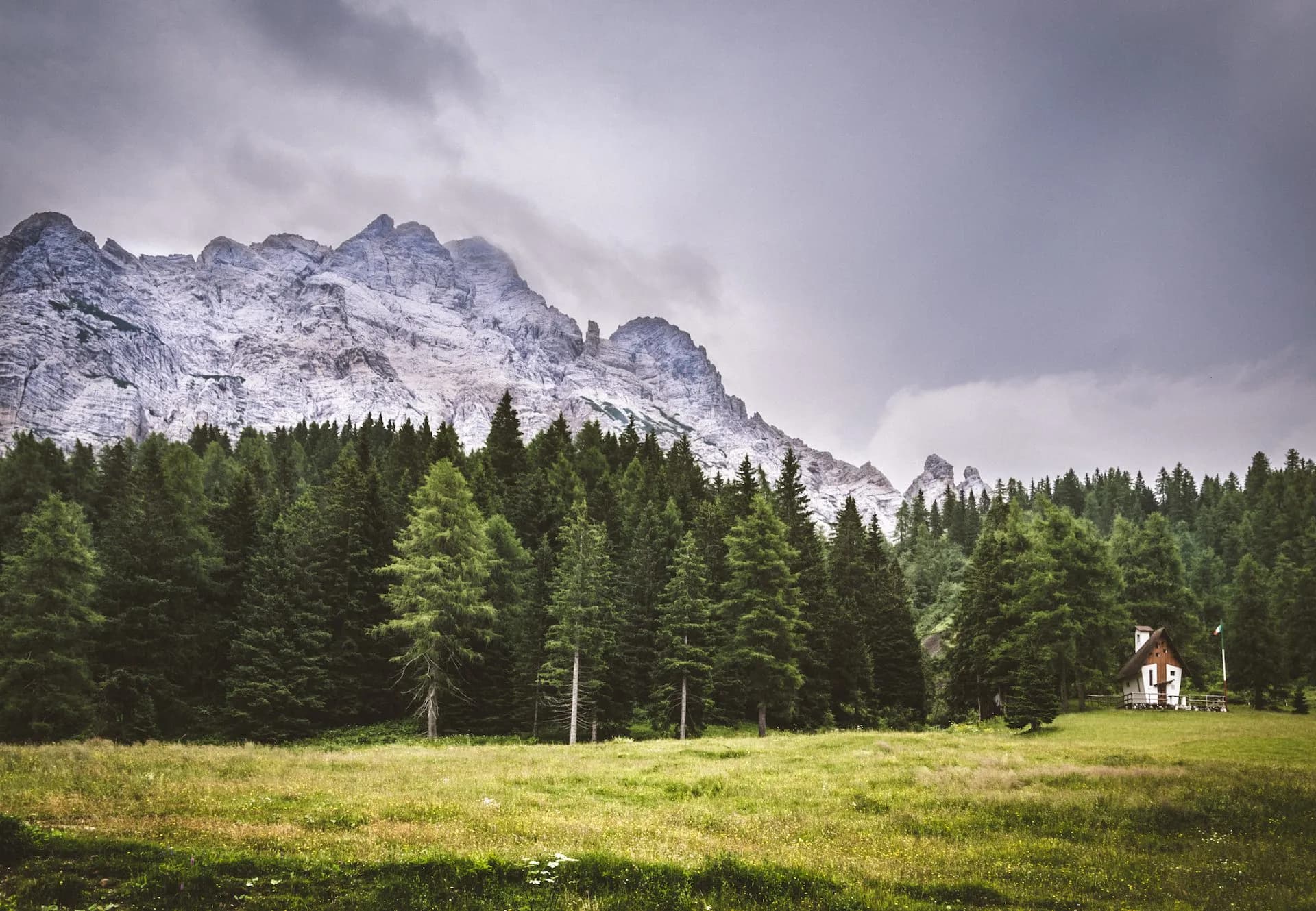 Mountain landscape with rocky peaks, dense pine forest, meadow, and small cabin near Passo Stulanza.