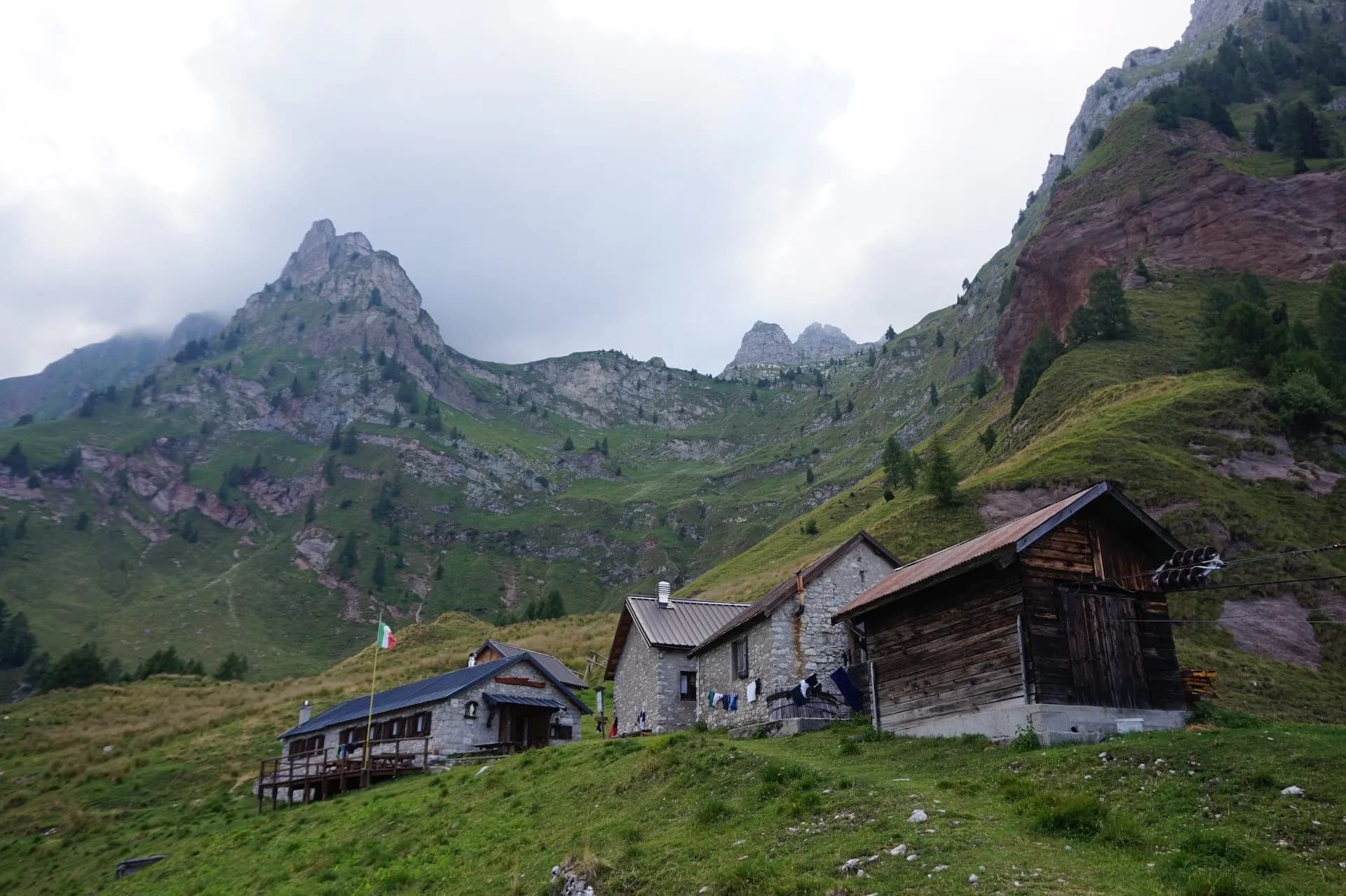 Rifugio Pian de Fontana mountain huts nestled on green slopes below rocky peaks under cloudy sky.