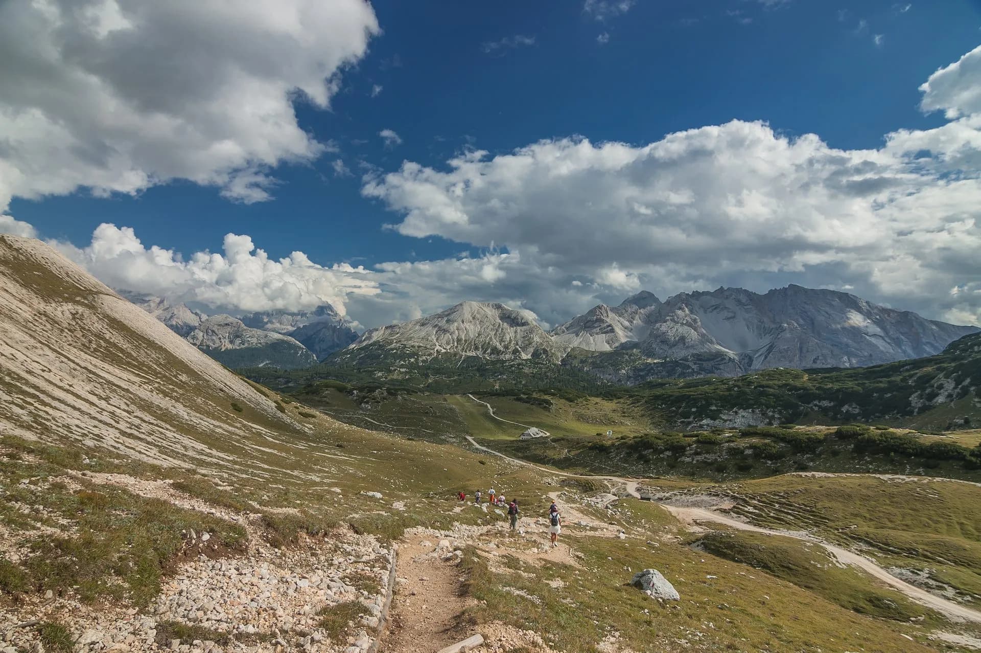 Hikers on rocky trail with green slopes and rugged Dolomite mountains under cloudy sky.