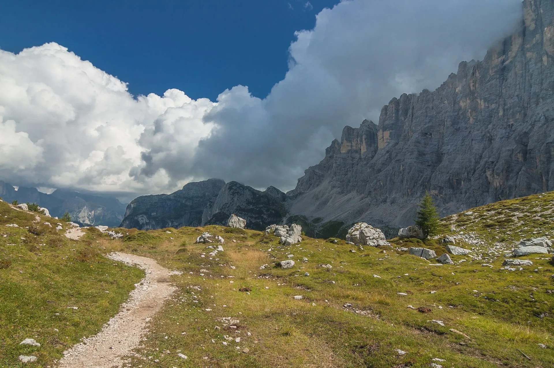 Hiking trail across grassy alpine meadow toward towering gray mountain cliffs under dramatic clouds.