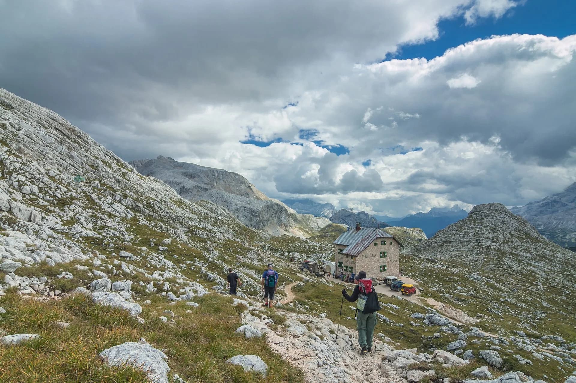 Hikers approaching Rifugio Biella mountain hut among rocky slopes under a dramatic cloudy sky.