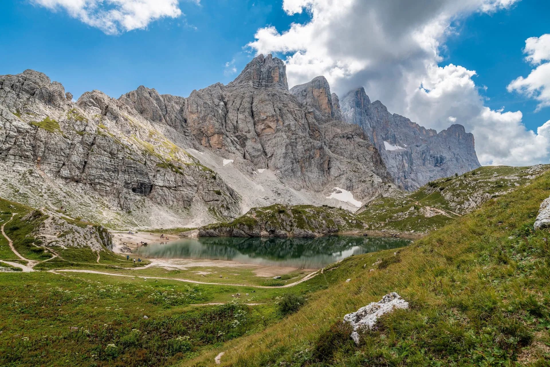 Alpine lake reflecting jagged gray mountains under a partly cloudy blue sky with hikers nearby.